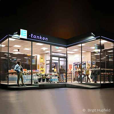 Außenansicht einer beleuchteten, belebten Tankstelle mit großen Glasfenstern bei Nacht.