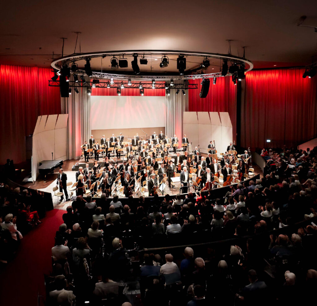 Ein großes Orchester spielt in einem Konzertsaal mit roter Beleuchtung.