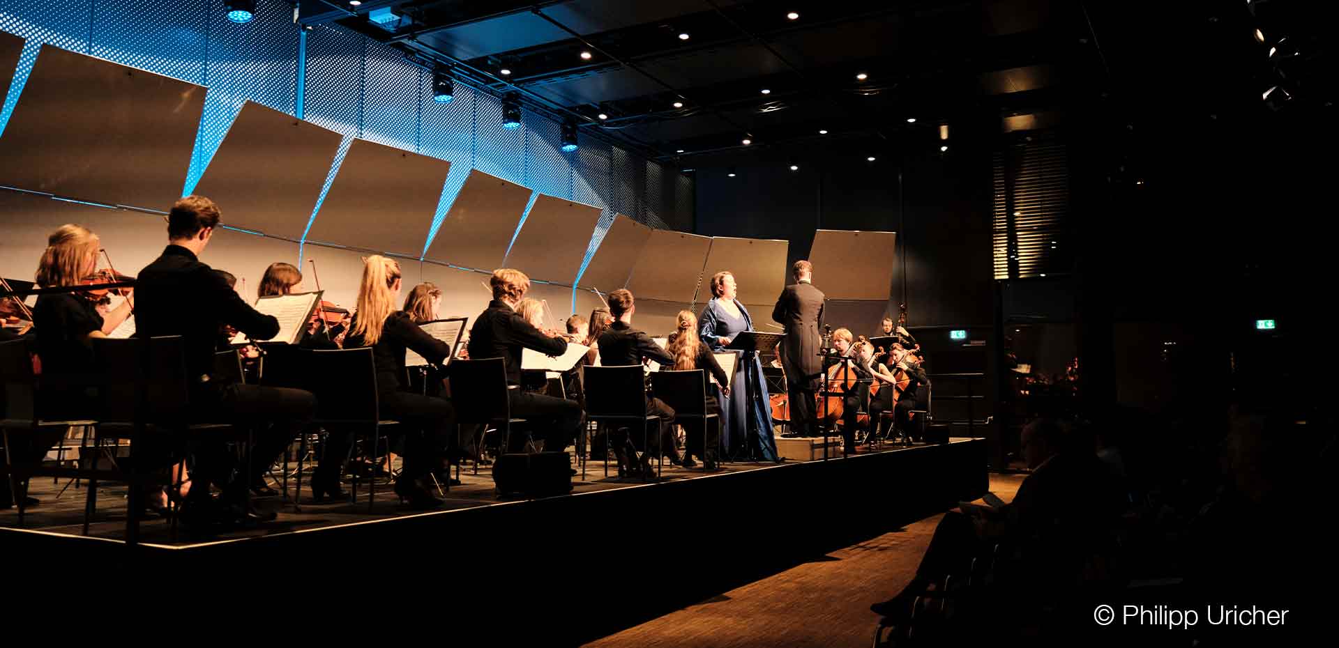 Ein Orchester spielt auf einer Bühne, zwei Sänger vorne in eleganter Kleidung