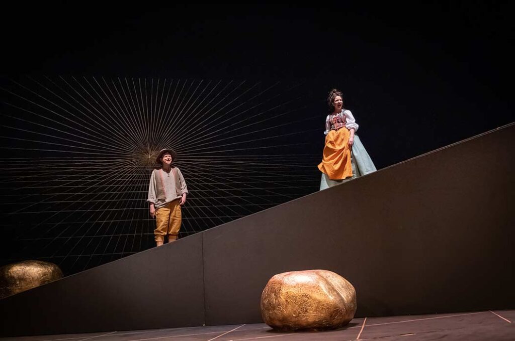 Zwei Schauspieler mit ernsten Mienen auf schräger Bühne, goldene Steine im Vordergrund