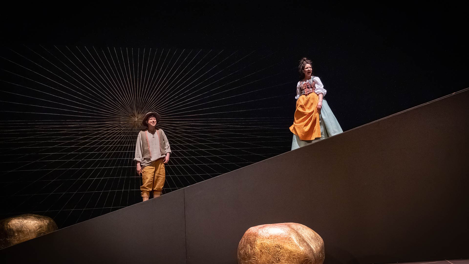 Zwei Menschen auf einer schrägen Bühne, beide farbenfroh gekleidet und lächelnd