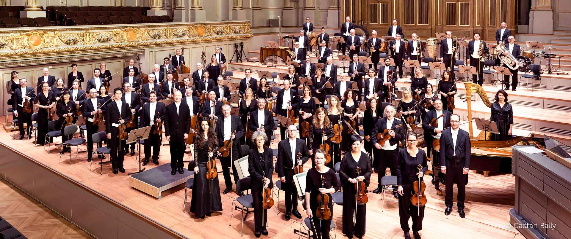Ein großes Orchester mit vielen Musikern in schwarzer Kleidung auf einer Bühne, einige stehen mit Instrumenten und wirken konzentriert
